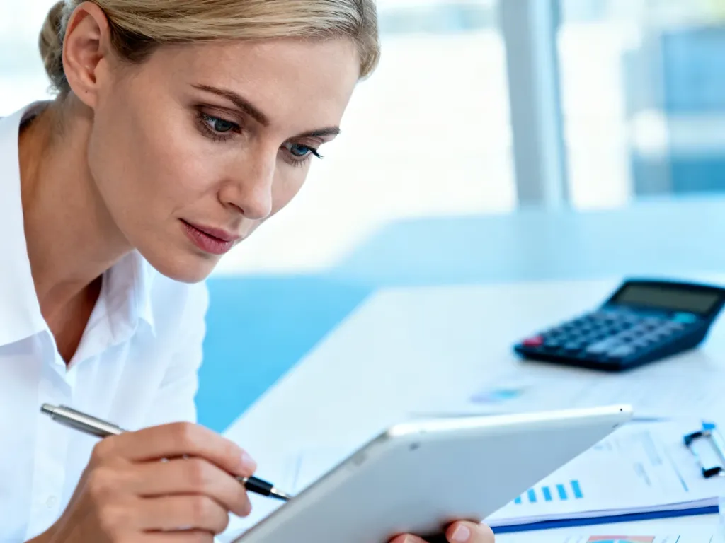 Kobieta z uwagą analizuje dane finansowe na tablecie w nowoczesnym biurze, co ilustruje proces obliczania wynagrodzenia.